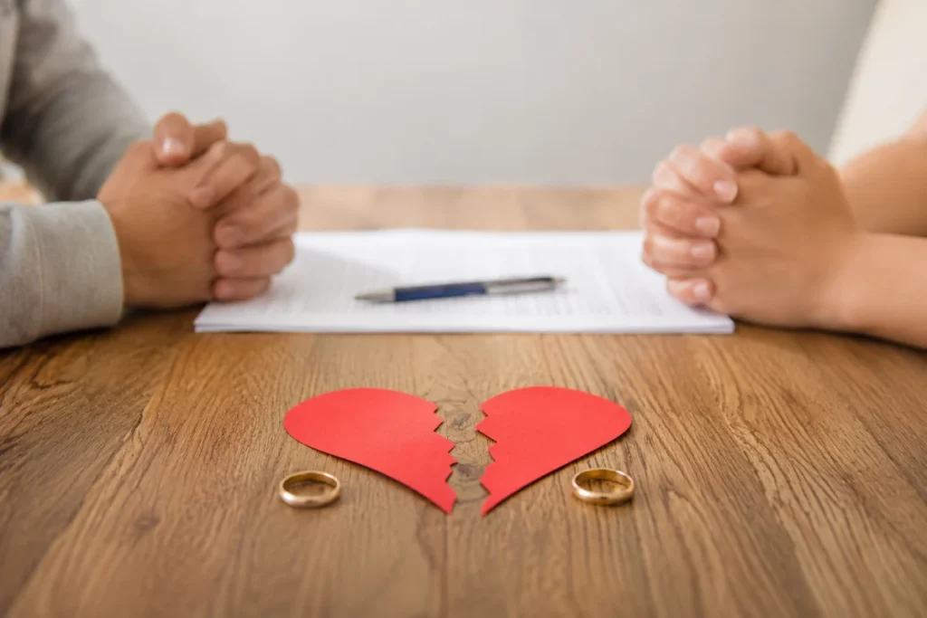Filing for divorce in Texas: a torn red paper heart and separated wedding rings on a wooden table between a man and woman.